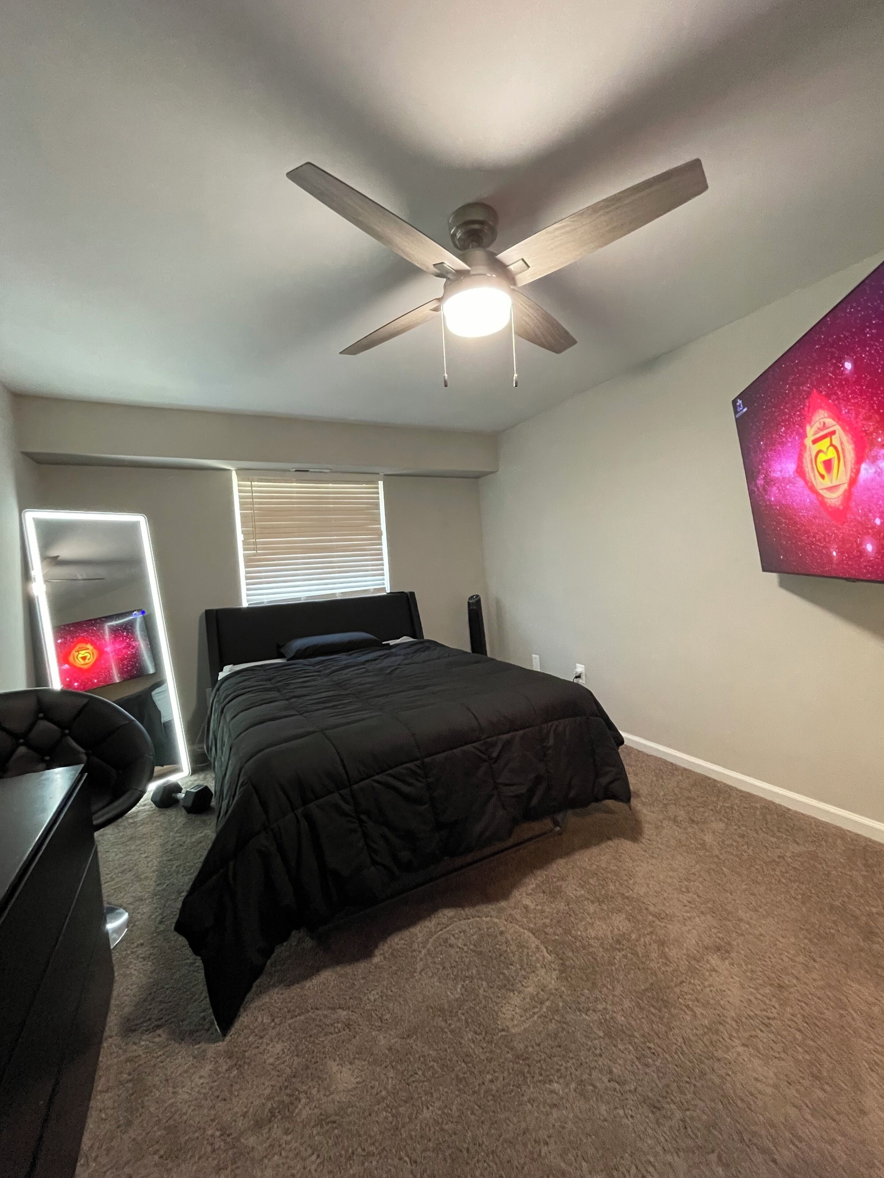Ein Schlafzimmer mit einem Bett, einem Deckenventilator, einem an der Wand befestigten Fernseher und einem Fenster mit Jalousien.
