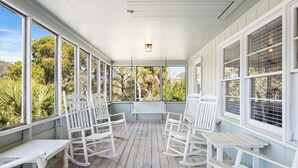 Une véranda couverte avec des fauteuils à bascule et des tables blancs, de grandes fenêtres et une vue sur la verdure.