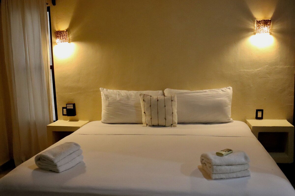 Quarto de hotel com cama, duas mesinhas de cabeceira e duas luzes fixadas na parede.