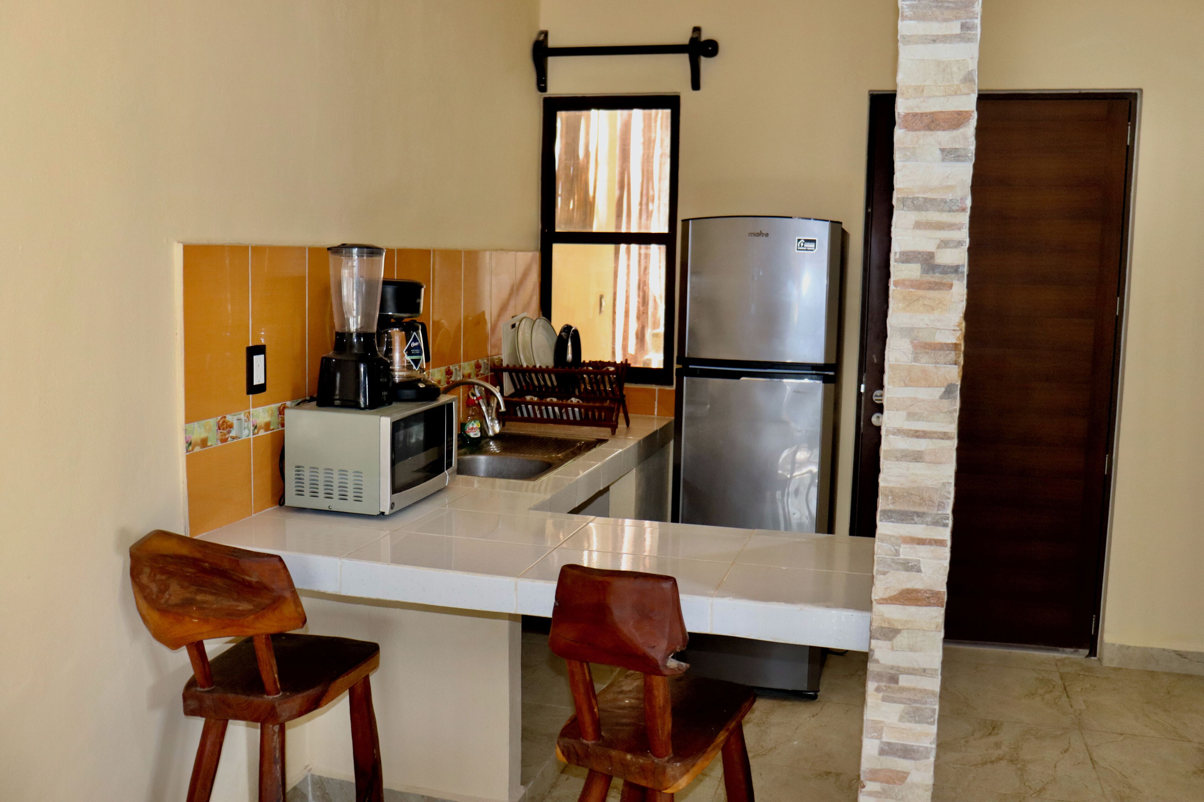 A compact kitchen with a refrigerator, microwave, and coffee maker. There are two wooden chairs and a small island with a sink.