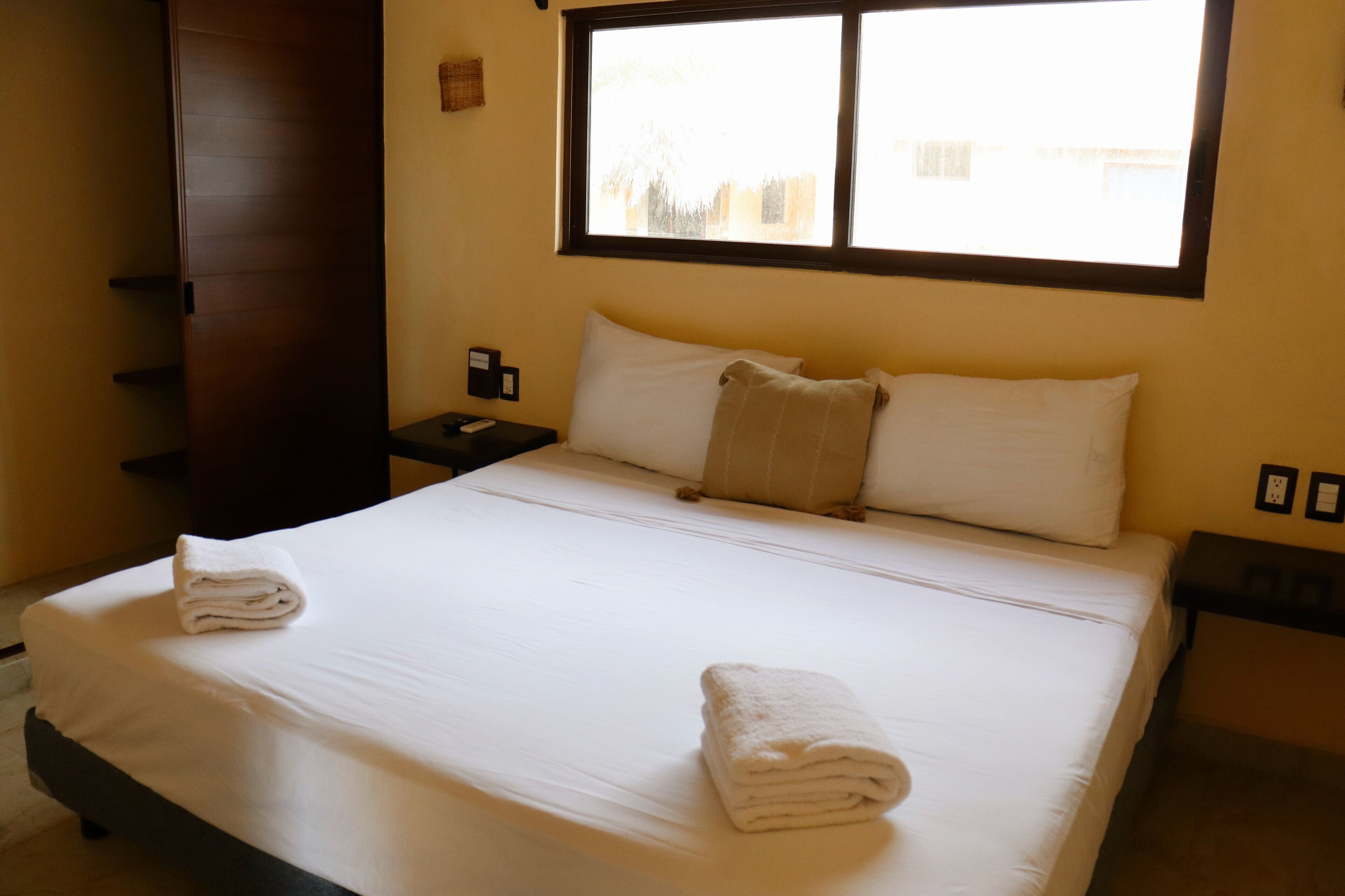 A neatly made bed with white linens and two pillows, a wooden wardrobe, a nightstand with a phone, and a window with a view of buildings.