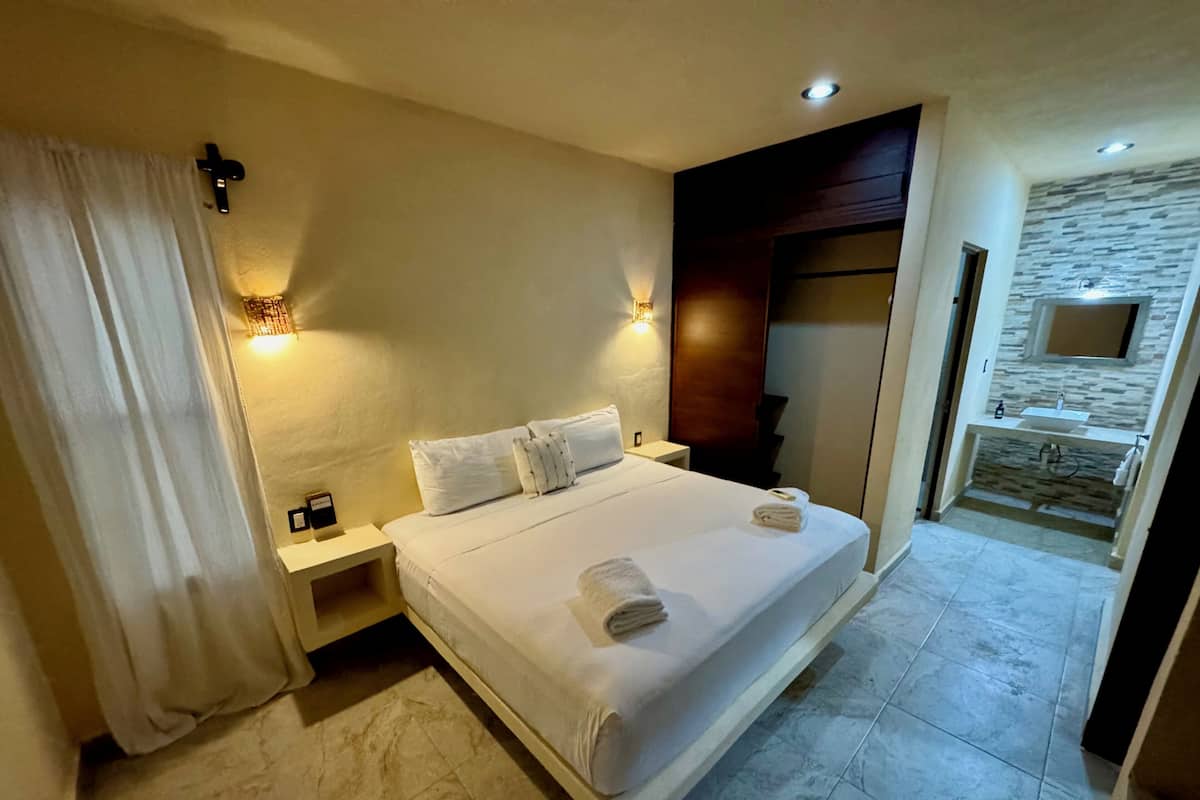 Una habitación de hotel con cama, mesitas de noche, una lámpara fijada a la pared, un armario de madera y un baño con pared de piedra y lavamanos.