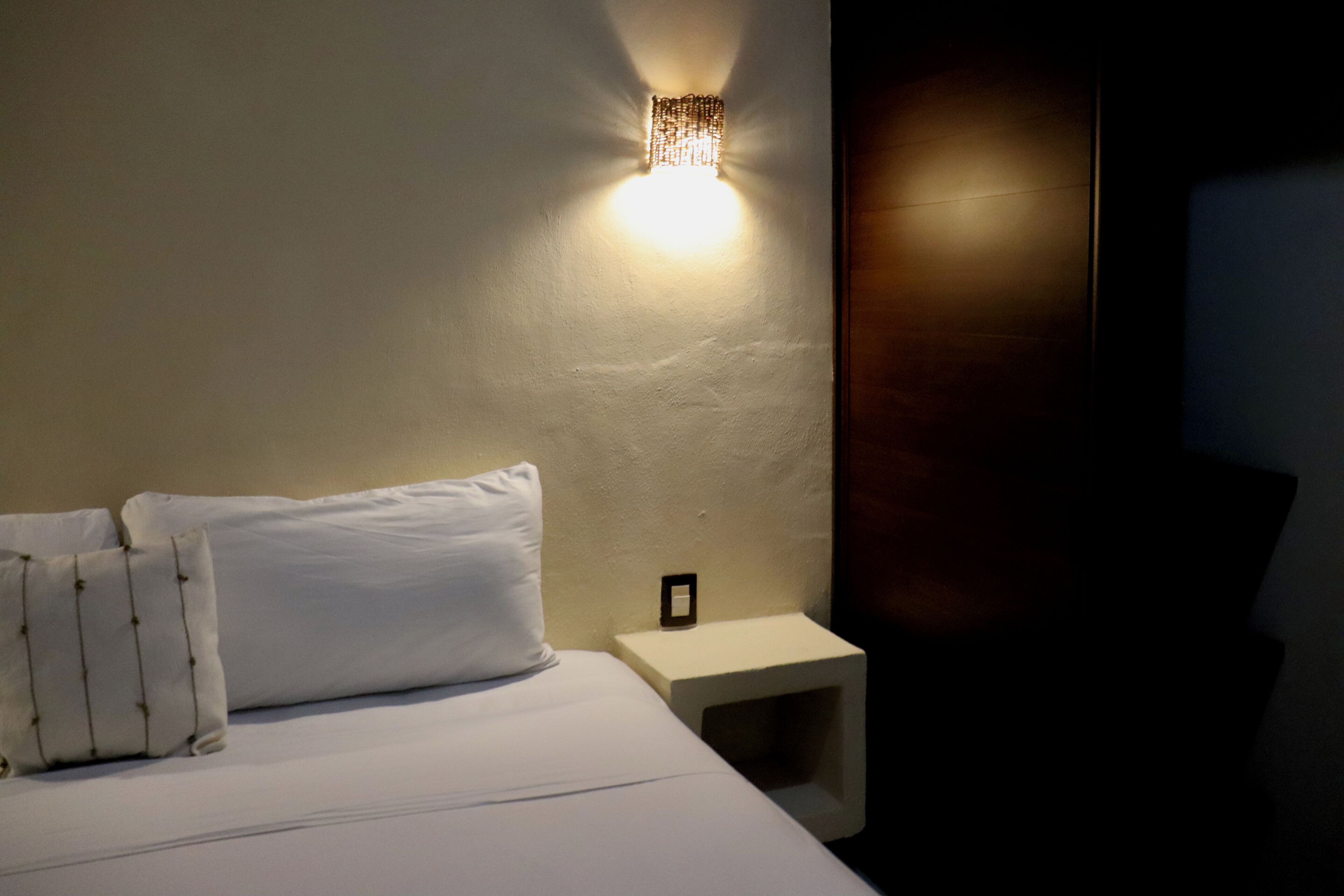 A hotel room with a bed, pillows, a nightstand, and a wall-mounted light fixture.
