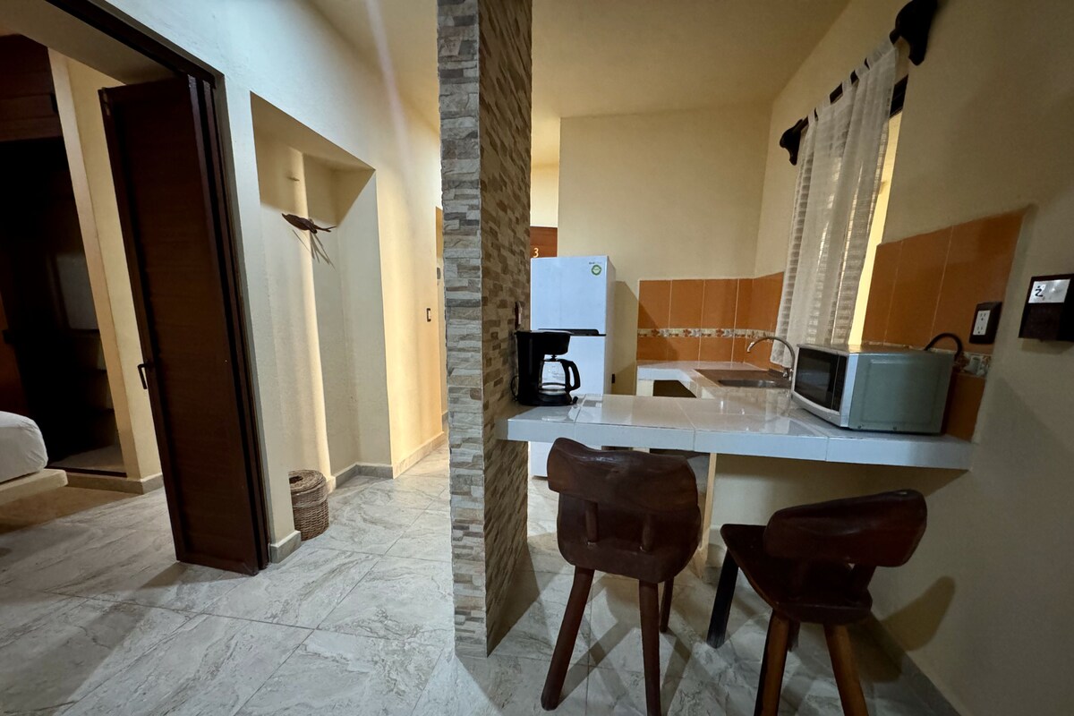 A compact kitchen with a microwave, coffee maker, and a sink. There are two wooden stools and a small wicker basket.