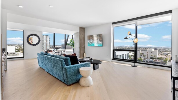 Ein geräumiges Wohnzimmer mit einem blauen Sofa, einem runden Couchtisch und einem großen Fenster mit Blick auf die Stadt.