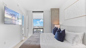 Ein modernes Schlafzimmer mit einem großen Bett, einem Flachbildfernseher, Blick auf die Stadt und einem gerahmten abstrakten Gemälde.