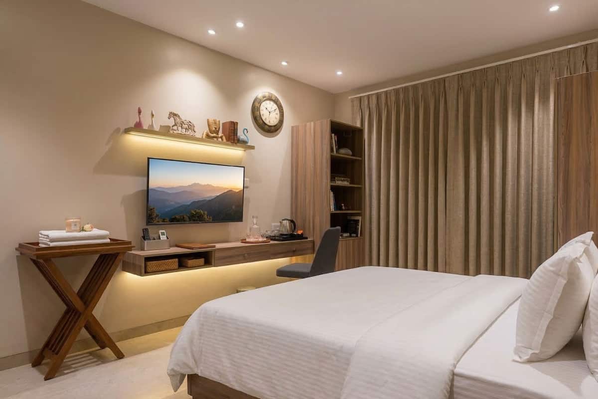 A modern hotel bedroom with a bed, a desk, a TV, and a bookshelf.