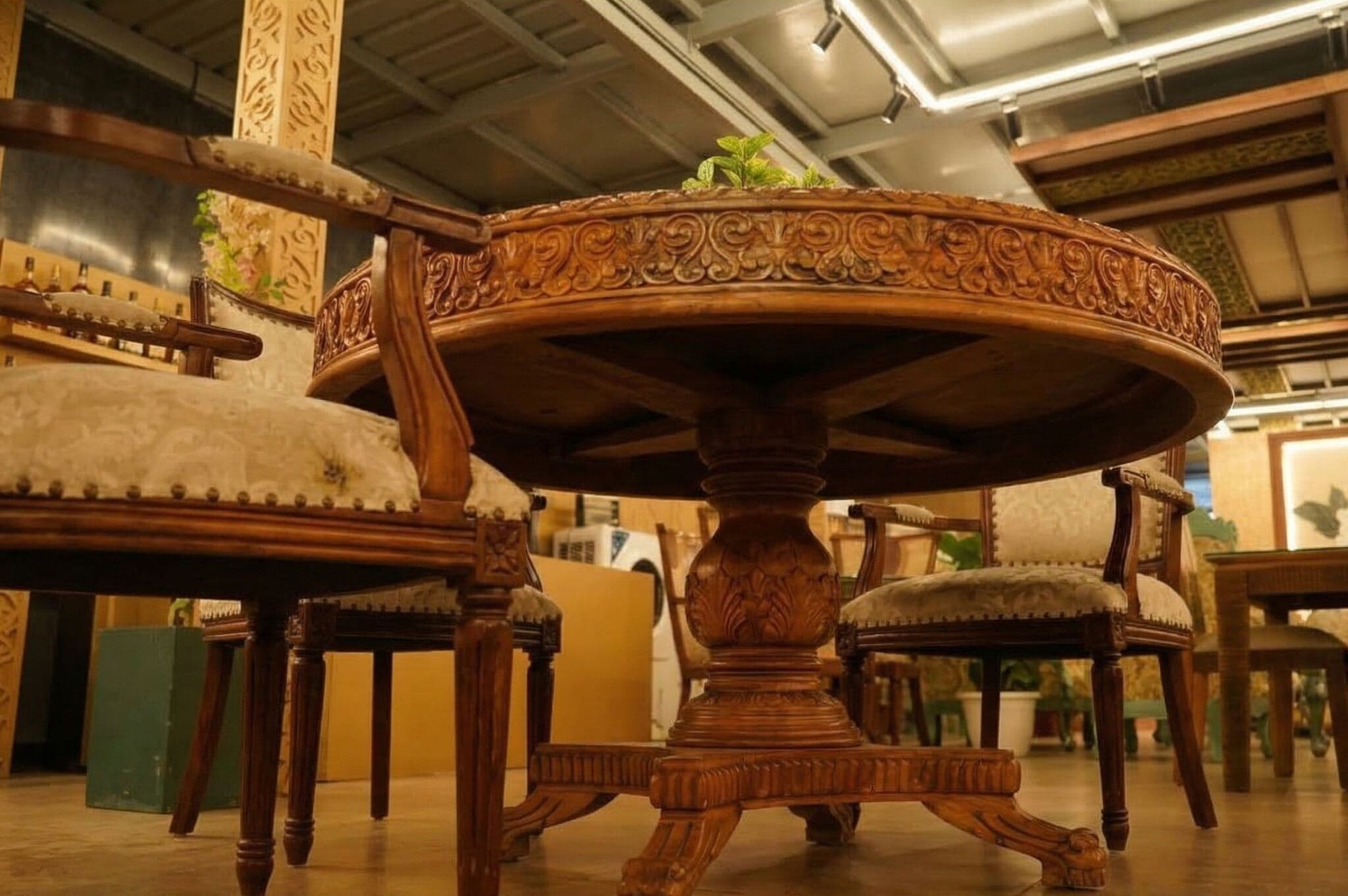 A round wooden table with intricate carvings, surrounded by upholstered chairs with a floral pattern.