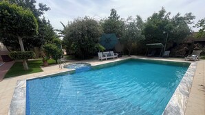 Piscina rectangular con borde de azulejos, rodeada por un patio empedrado y adyacente a una zona verde con árboles y un edificio.