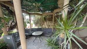 Un espacio de estar al aire libre cubierto con una mesa y bancos, rodeado de plantas en macetas.