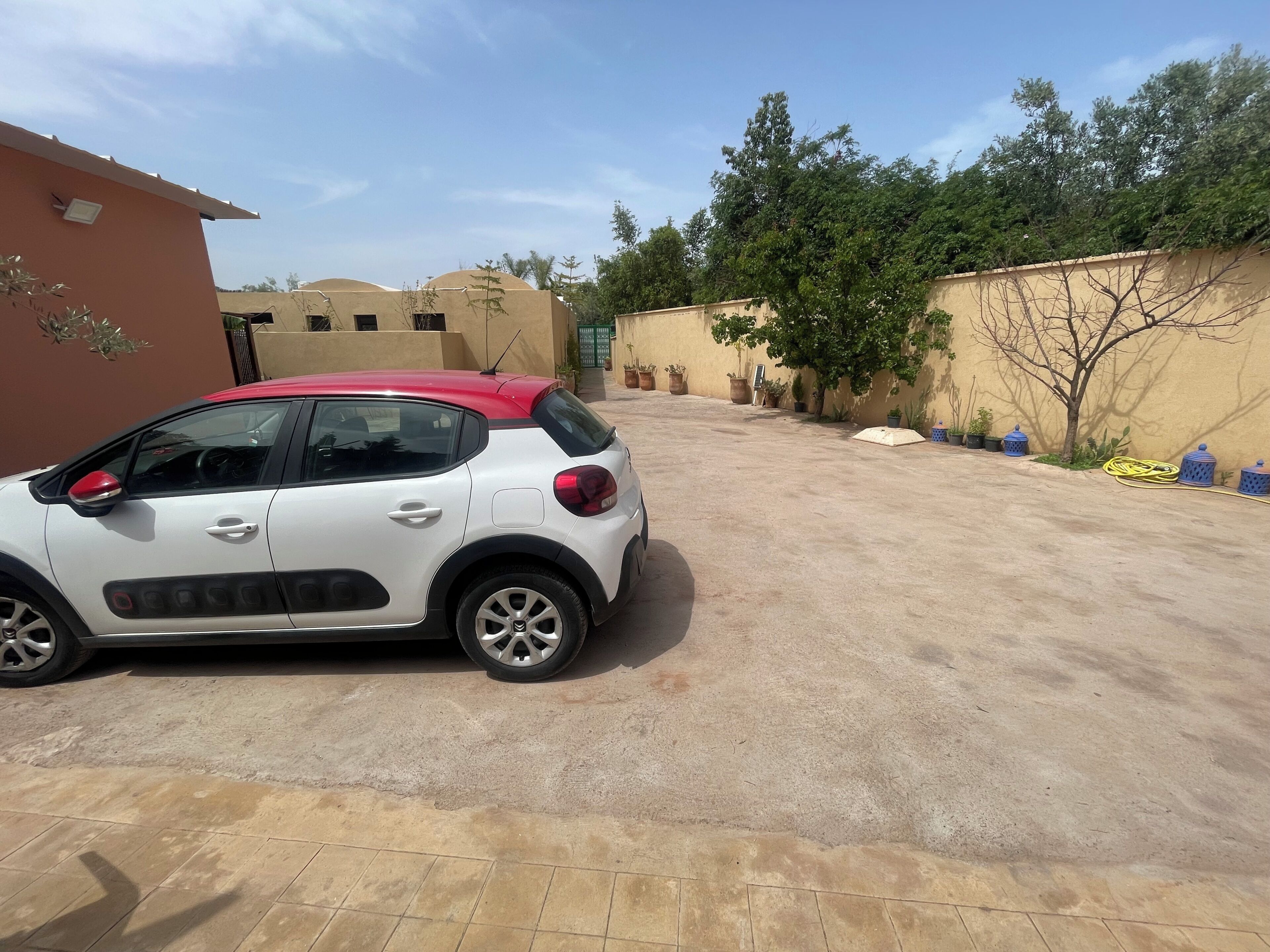 Une voiture blanche est garée dans une allée, avec un mur de couleur beige et des plantes en pot en arrière-plan.