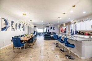 A modern kitchen with a central island, bar stools, and a dining area with a long table and chairs. - NEW!Racing Simulator|Game Room|Movie Room|Sleep 21 (Kissimmee)