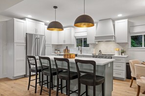 Une cuisine moderne avec des meubles blancs, un îlot central doté de tabourets, des appareils en acier inoxydable, un éclairage suspendu et une vue sur la verdure à l’extérieur.