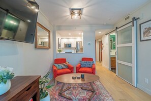 Ein modernes Wohnzimmer mit Fernseher, zwei roten Sessel, einem Couchtisch und einer Küche im Hintergrund.