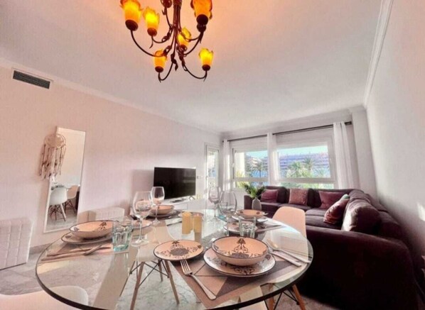 A modern living room with a dining table set for four, a sofa, a TV, and large windows. - Sea front apartment in Playa Rocio, Puerto Banus with swimming pool and parking (Marbella)