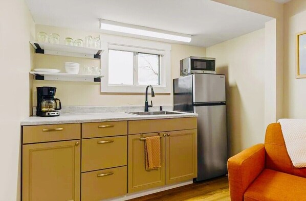A compact kitchen with a sink, a microwave, a refrigerator, and a coffee maker. There is a window above the sink and a chair in the corner.