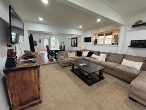 Ein geräumiges Wohnzimmer mit einem Ecksofa, einem Couchtisch und einer Holzkabine. Im Hintergrund ist eine Essecke mit Tisch und Stühlen zu sehen.