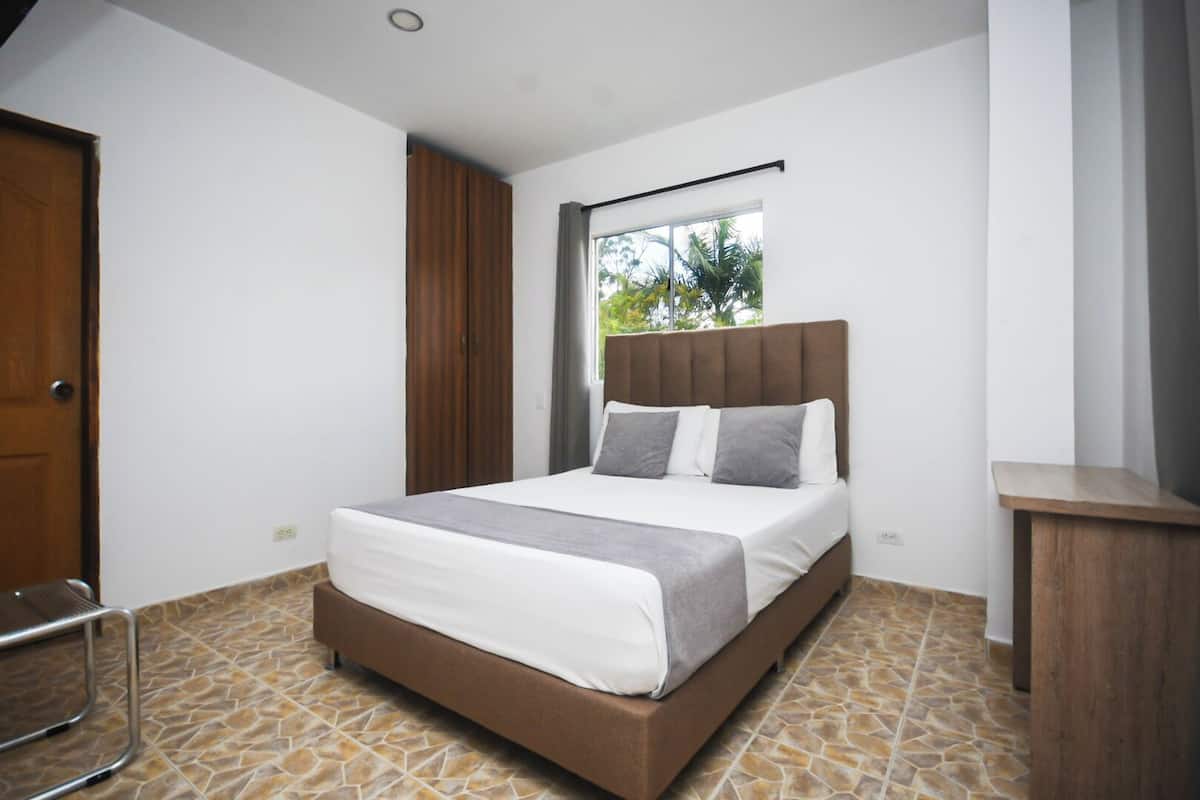 A bedroom with a bed, a window with curtains, a door, and a small wooden table.