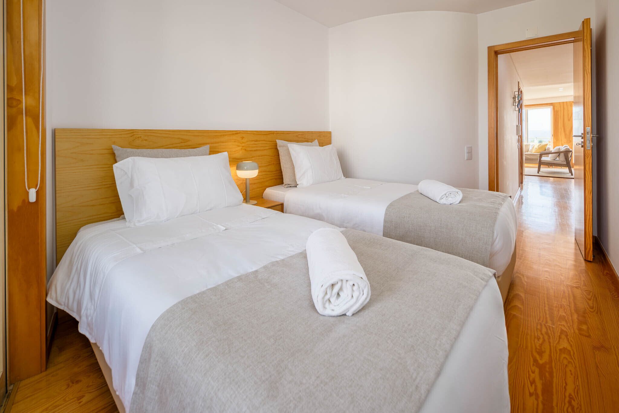 A hotel room with two beds, wooden headboards, a nightstand with a lamp, and a wooden floor.