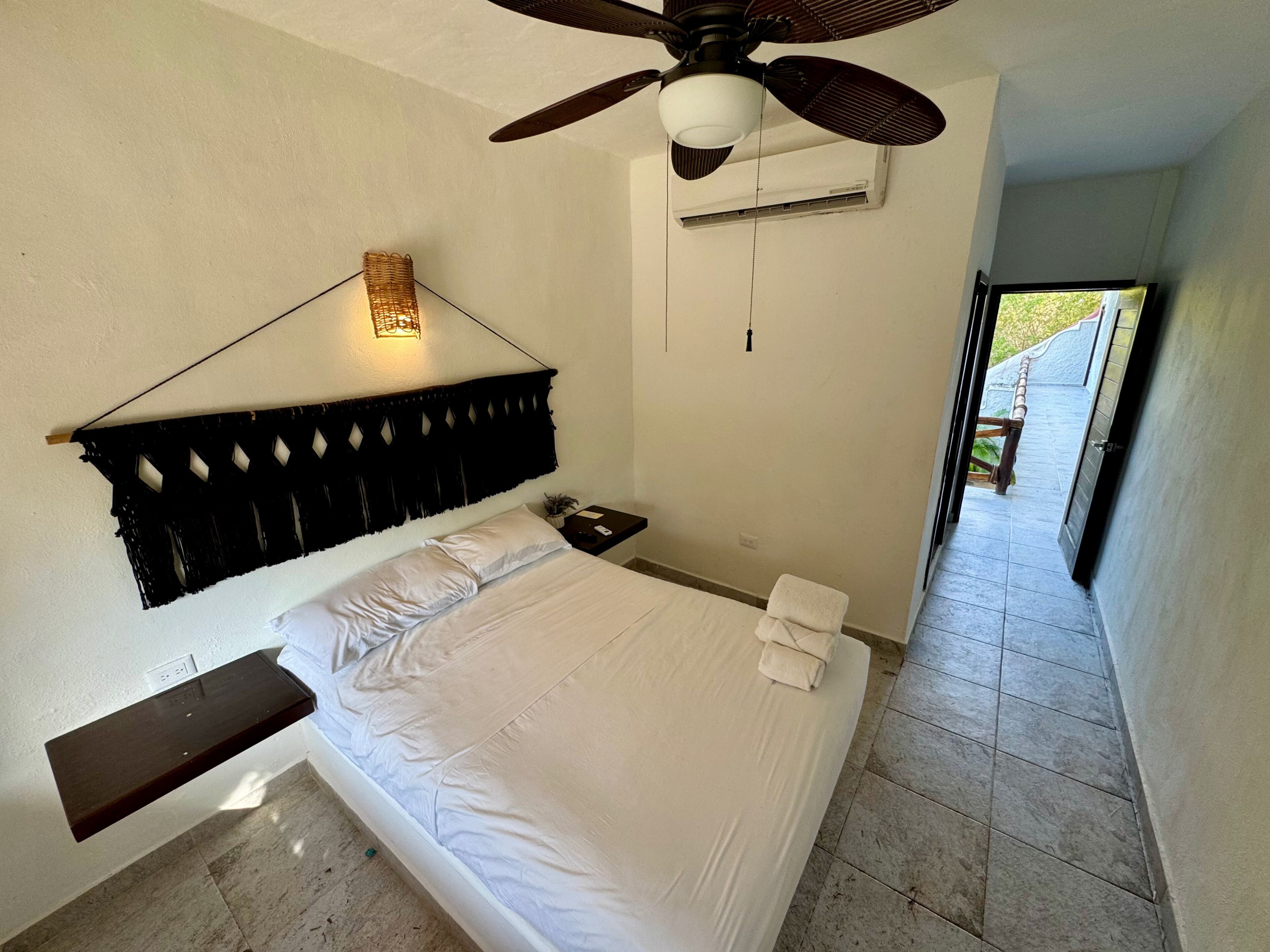Une chambre à coucher comprenant un lit, une table de chevet, un ventilateur de plafond, un appareil de climatisation et une vue sur l’extérieur.