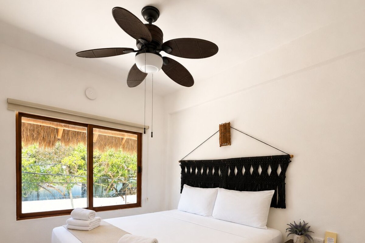 A bedroom with a bed, a ceiling fan, a window with a view, and a bedside table with a plant.