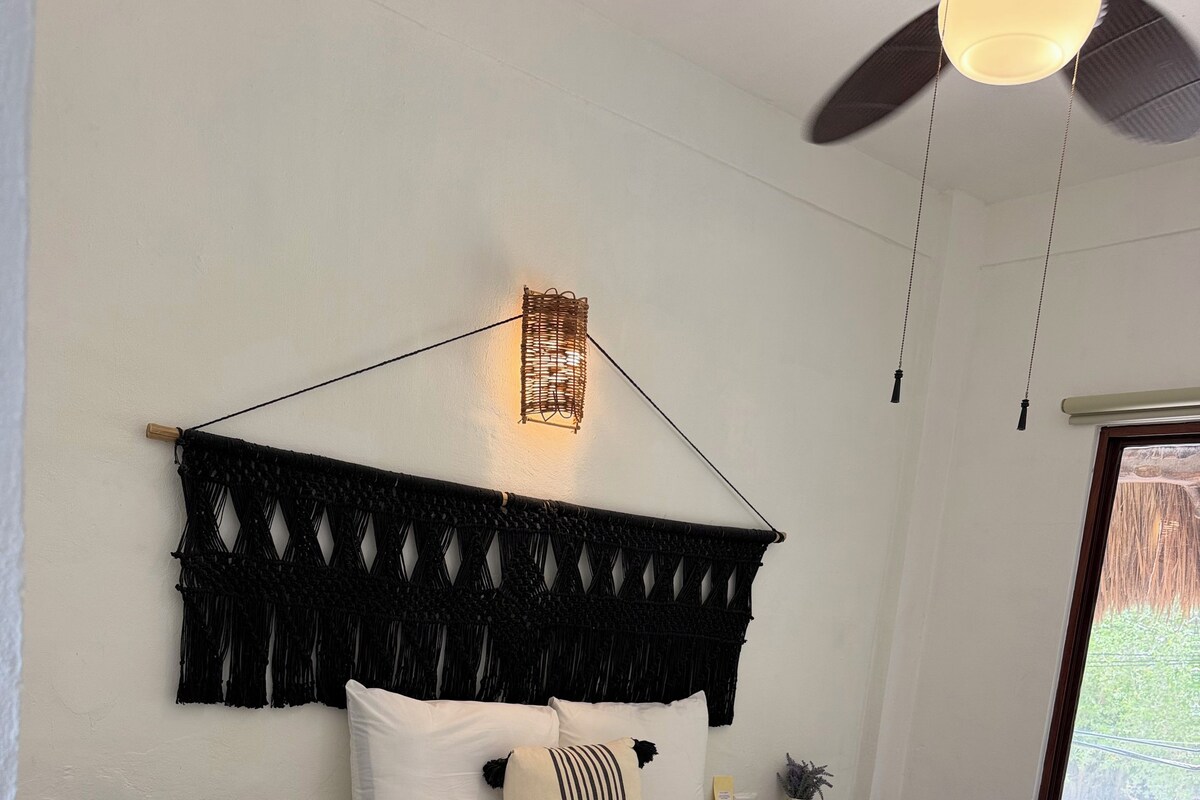 A neatly made bed with white linens, a black macrame headboard, and a bedside table with a lamp.