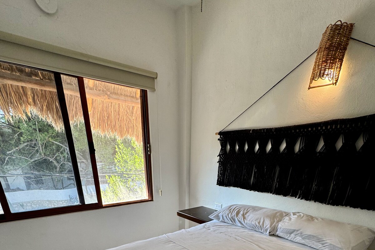A bedroom with a bed, a bedside table, a wall-mounted light fixture, and a window with a view of greenery.