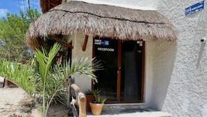 A thatched-roof building with a reception sign, a potted plant, and a blue sign indicating a location. - Paseo Kuka by ÓOL Hotels (Isla Holbox)