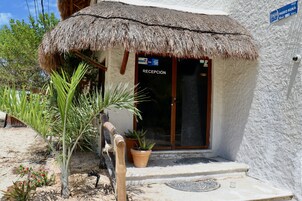Un edificio con techo de paja, un letrero de recepción, una planta en maceta y una señal azul que indica una ubicación.