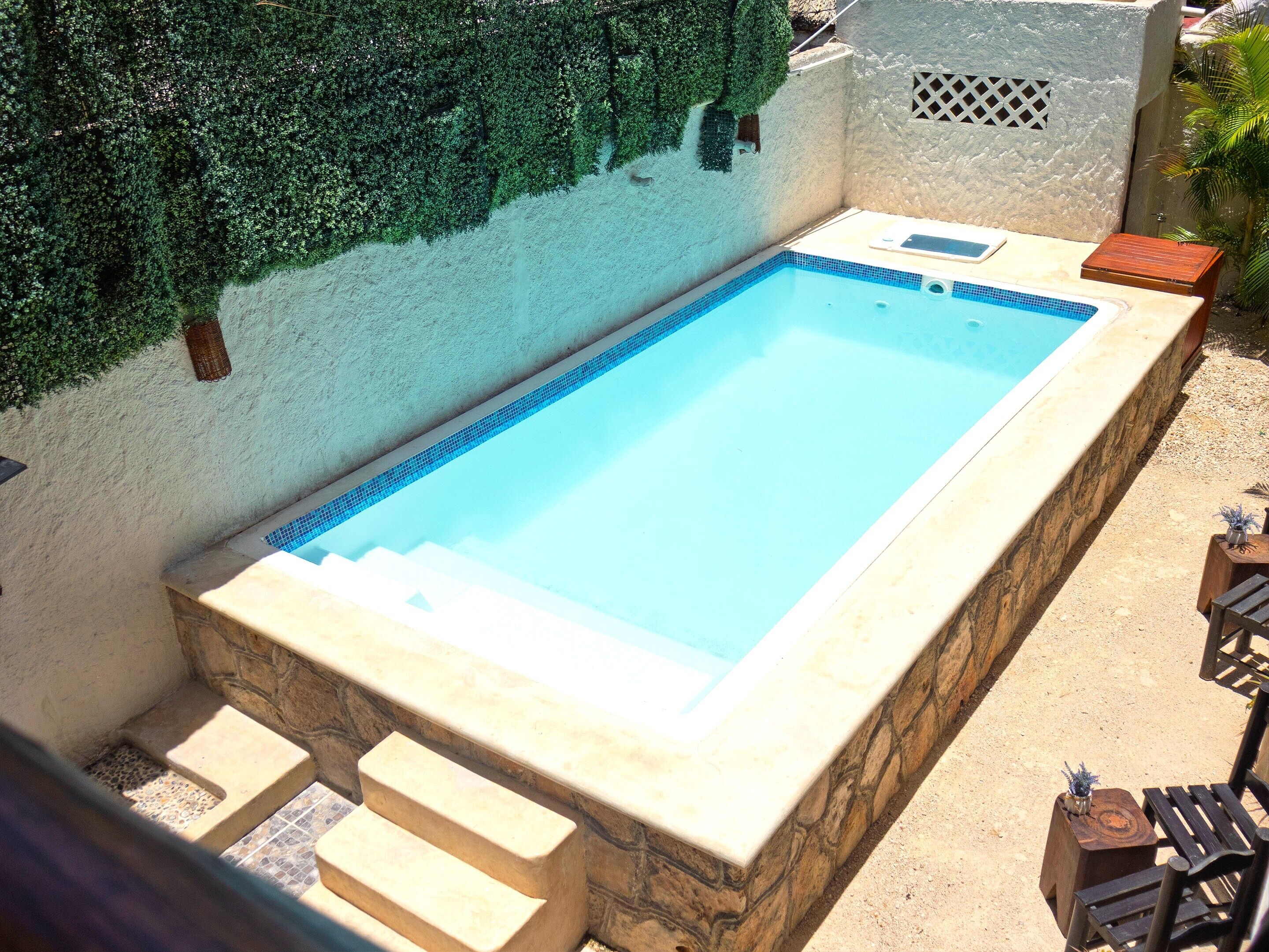 A rectangular swimming pool with a stone border, steps leading into the water, and a small wooden table with a plant beside it.
