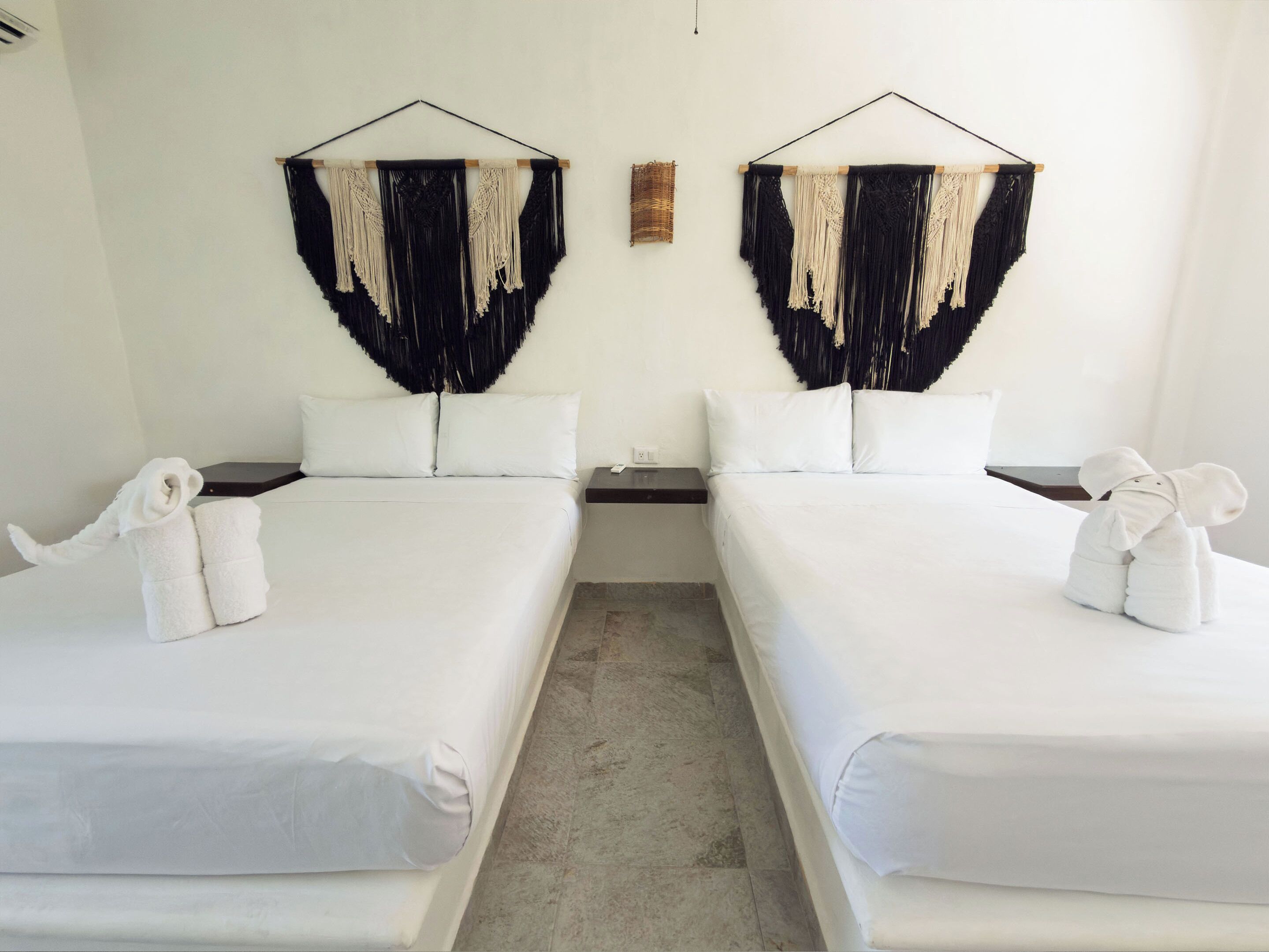 Habitación de hotel con dos camas, ropa de cama blanca y adornos de macramé en las paredes.