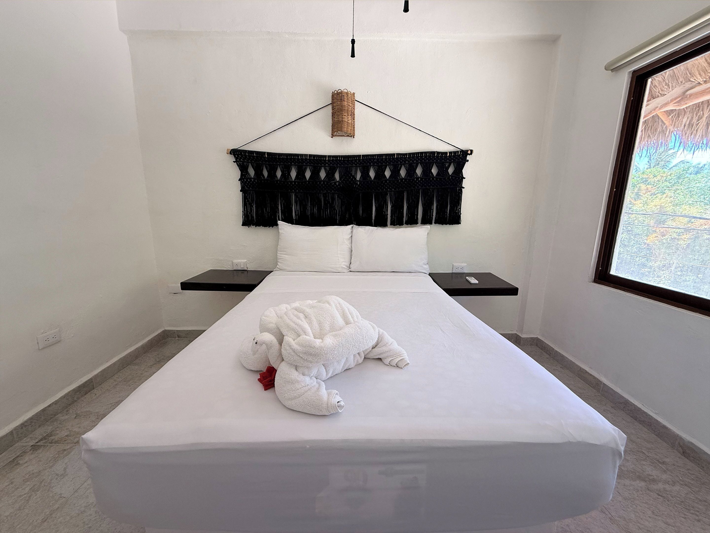 A bed with a white blanket, a white towel, and a white stuffed animal. There is a black headboard with a woven hanging decoration.