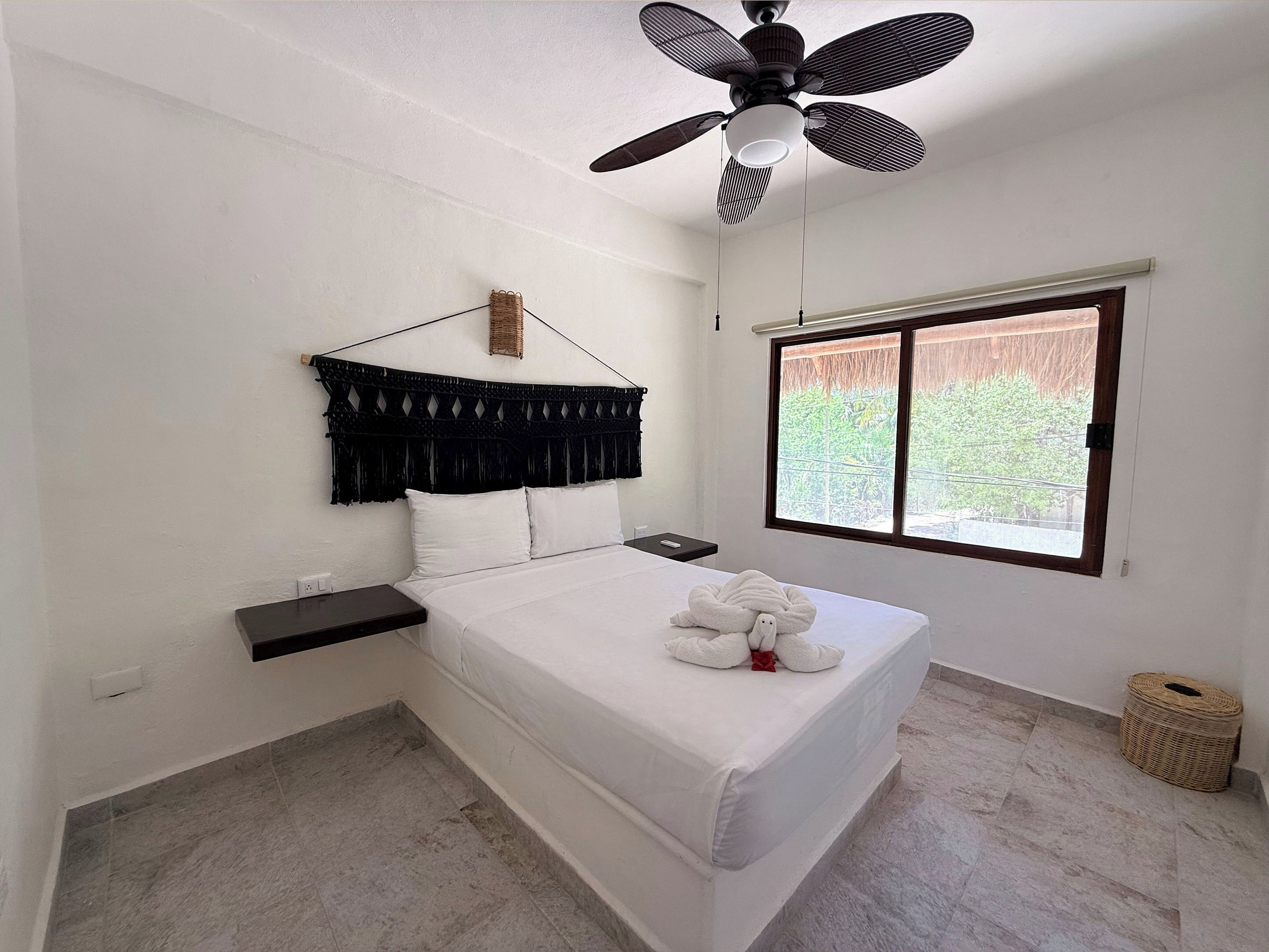 A bedroom with a bed, a ceiling fan, a window, and a small bedside table.