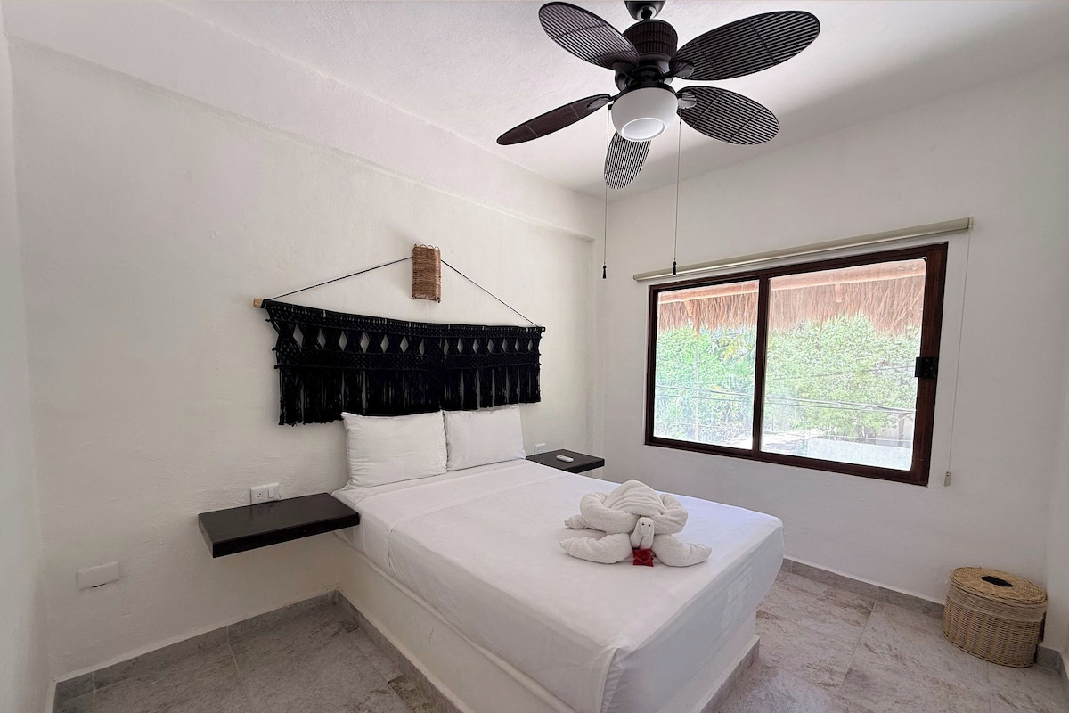 A bedroom with a bed, a ceiling fan, a window, and a small bedside table.