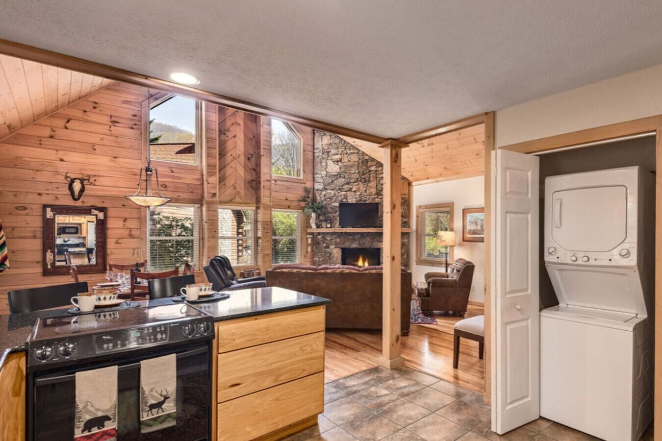 A rustic cabin interior with a wooden dining area, stone fireplace, and a large window overlooking a forest.