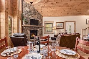 A cozy interior with a stone fireplace, wooden walls, and a dining area set for a meal. - Peace By The Creek-Hot Tub, Easy Access, Dogs Okay (Maggie Valley)