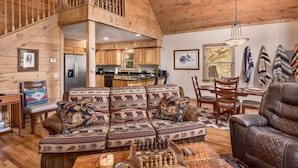 Ein gemütliches Cabin-Interieur mit einer Holztreppe, einer Couch mit gemusterten Kissen, einem Ledersessel und einem Schachbrett.