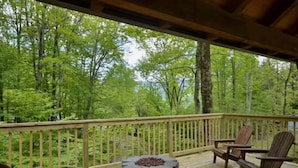 Eine Holzterrasse mit Adirondack-Stühlen und einer Feuerstelle aus Stein, umgeben von bewaldetem Gelände.