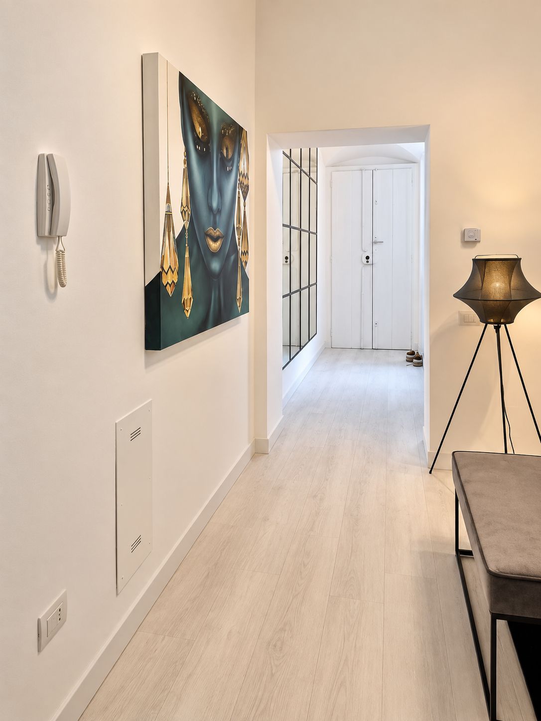 A hallway with a modern interior design, including a light-colored floor, a painting on the wall, a phone, and a lamp.