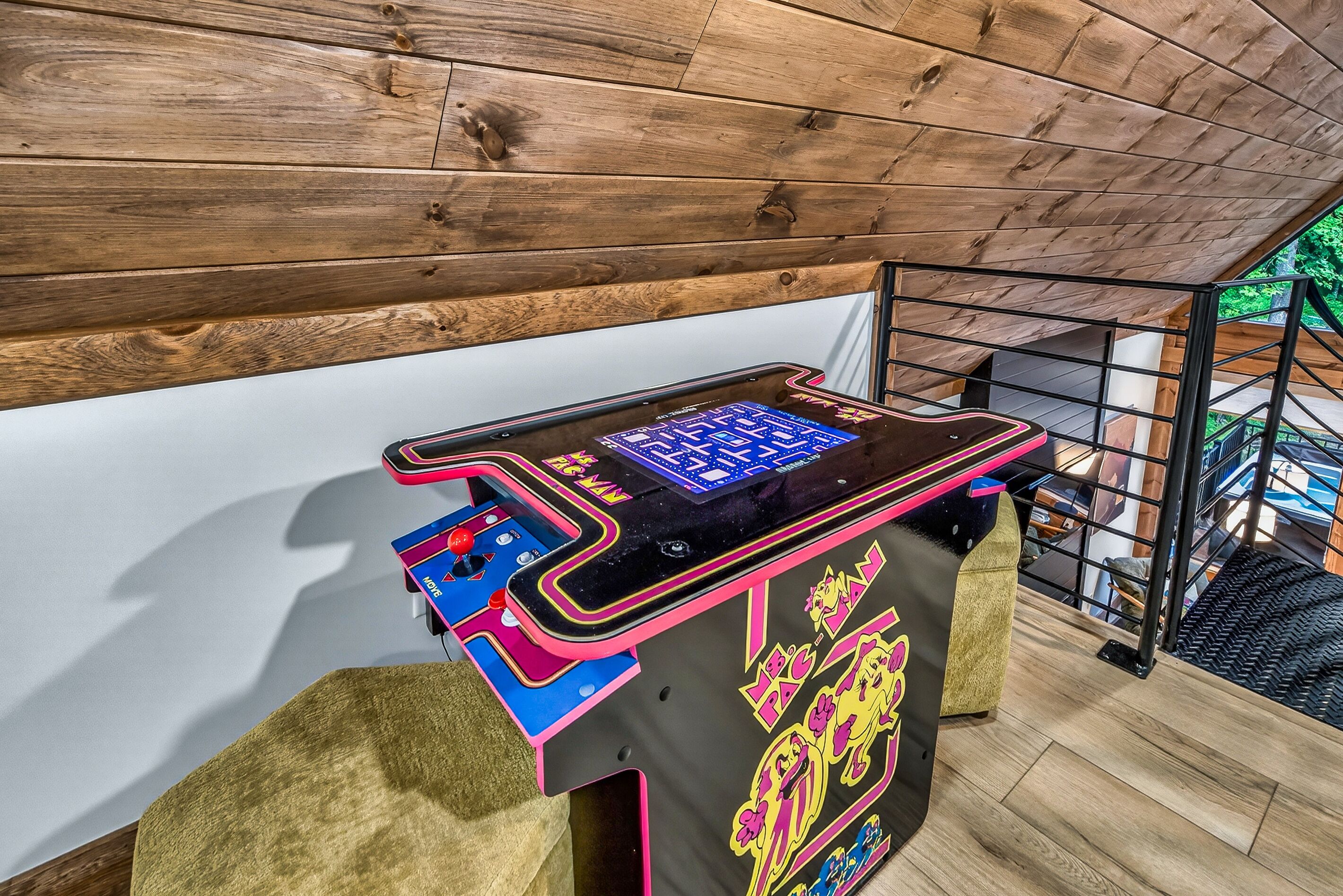 Una máquina de pinball en una habitación con techo y paredes de madera, una escalera y vistas a espacios verdes al exterior.