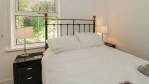 Ein Schlafzimmer mit Bett, einem Fenster mit Blick auf Grünflächen, einem Nachttisch mit Lampe und einer Kommode.