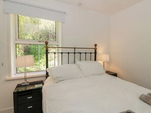 Ein Schlafzimmer mit Bett, einem Fenster mit Blick auf Grünflächen, einem Nachttisch mit Lampe und einer Kommode.