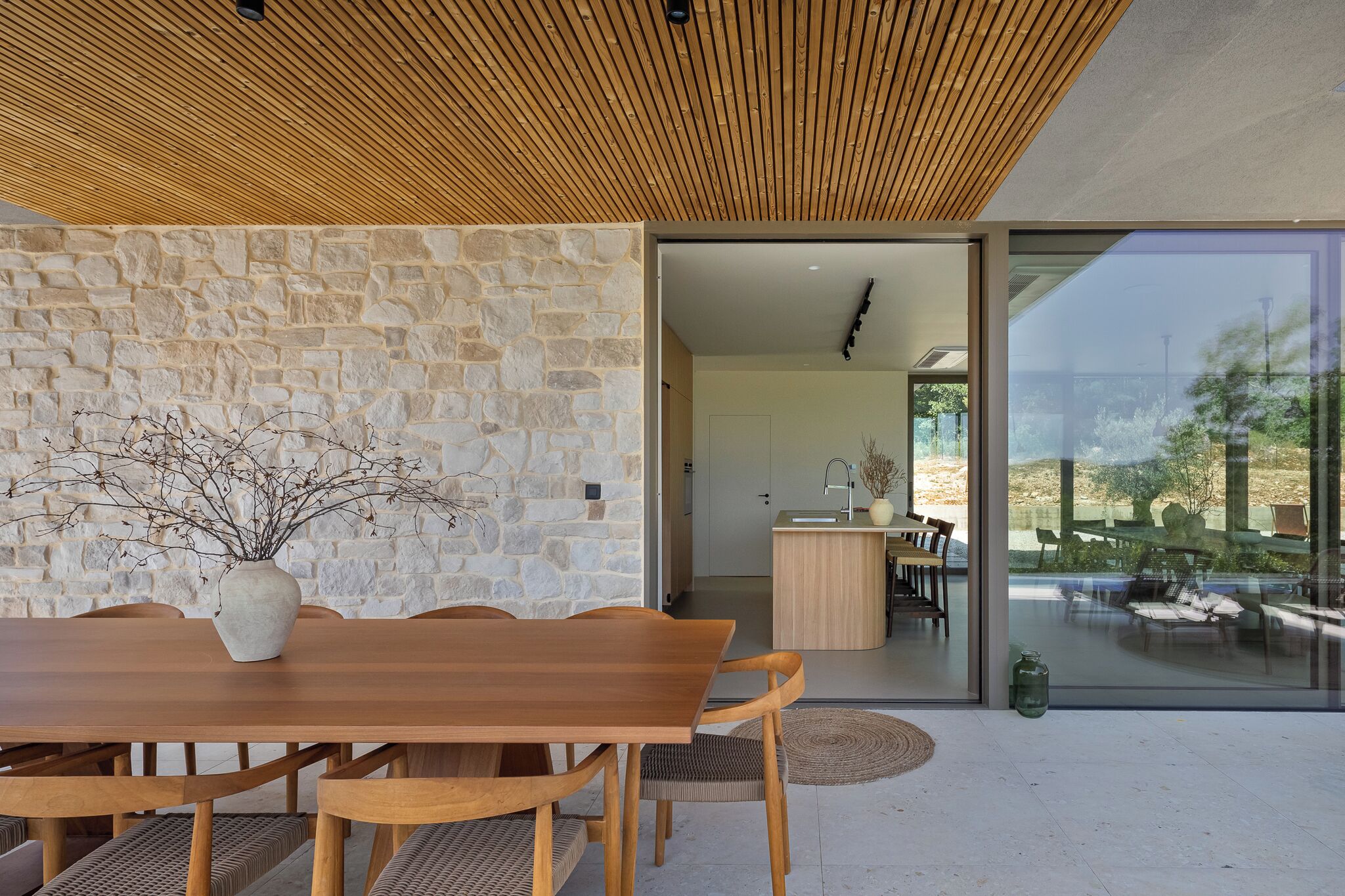 Uma sala de jantar com mesa e cadeiras de madeira, parede de pedra e vista através de portas de vidro para uma área externa com assentos.