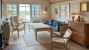 A cozy room with a wooden ceiling, a sofa with colorful pillows, a dining table, wicker chairs, a wooden dresser, and artwork on the walls.