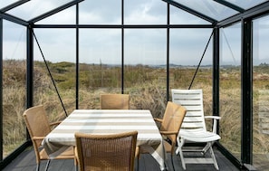 A glass-enclosed dining area with wicker chairs and a table, overlooking a natural landscape.