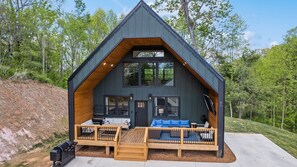 A cabin with a steep gable roof, a deck with seating, and a large window.