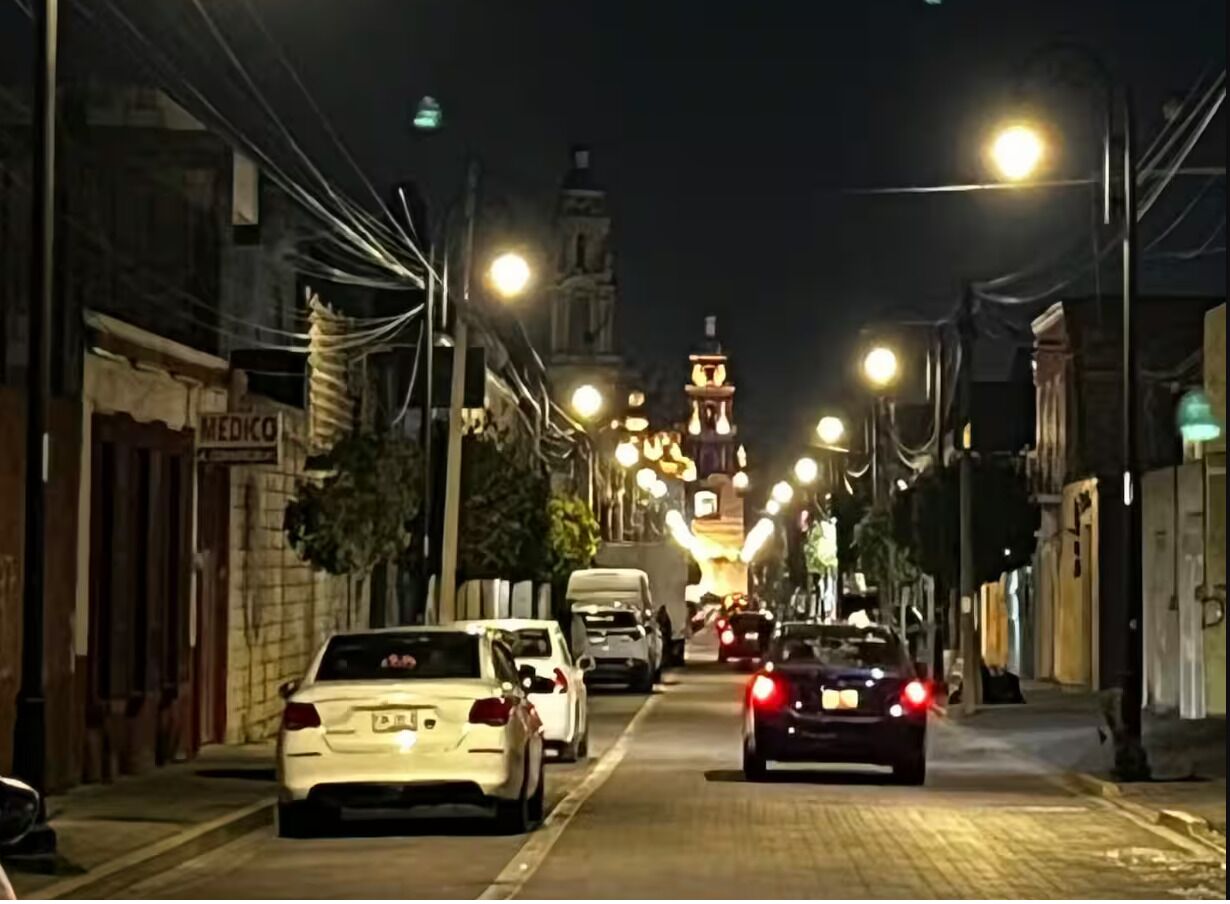 A nighttime street scene with cars parked on the side, buildings on either side, and a tower in the distance.