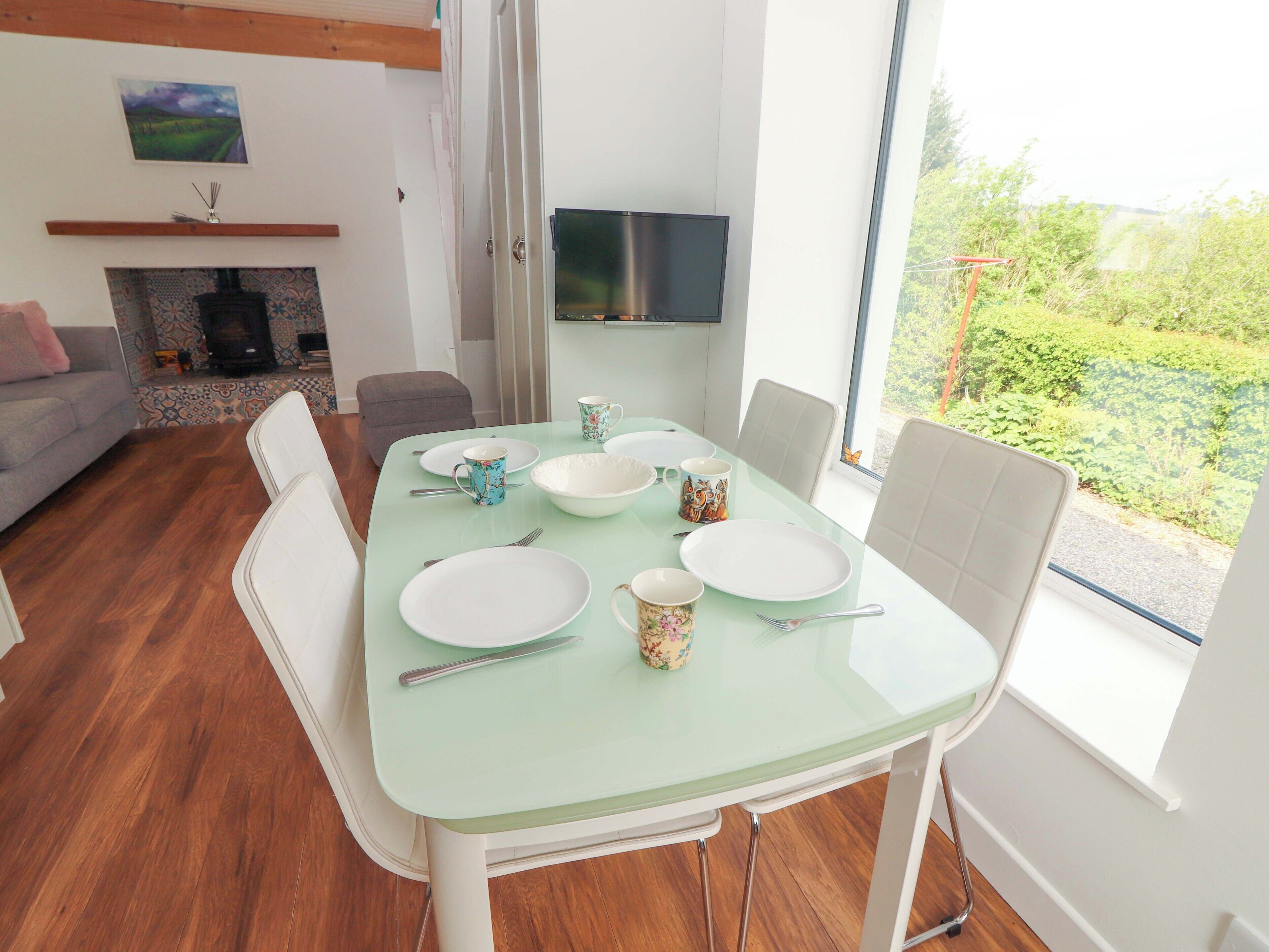 Een eetgedeelte met een glazen tafel, witte stoelen en een aan de muur bevestigde televisie.