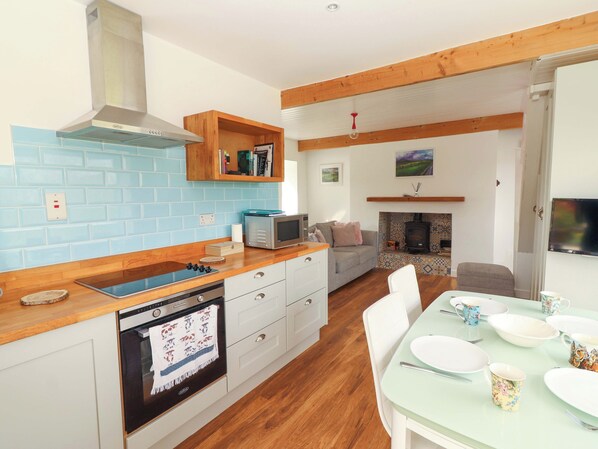 A modern kitchen with a dishwasher, oven, and wooden cabinets. There is a dining area with a table set for four, and a living room with a sofa and fireplace.