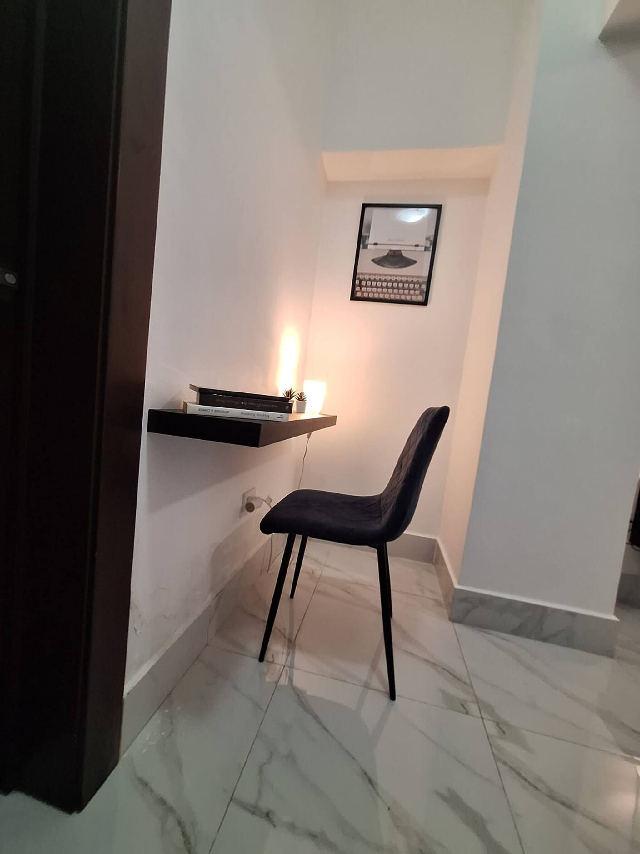 Una habitación pequeña y bien iluminada, con una silla de color oscuro, una estantería con una lámpara y un cuadro en la pared.